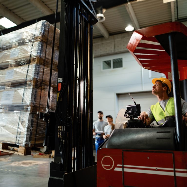 Forklift operator loading cargo while working in a warehouse. His colleagues are in the background.